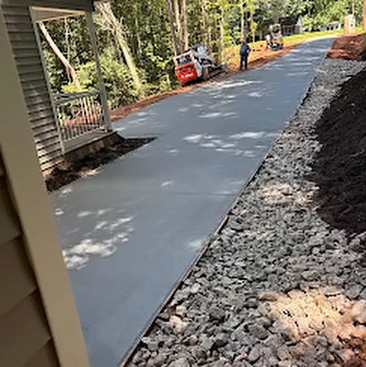 A finished concrete driveway on a wooded lot with a gravel edge